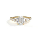 A yellow gold diamond ring featuring a large emerald-cut center stone flanked by smaller round and baguette-cut diamonds, set on a clean white background with a soft shadow.