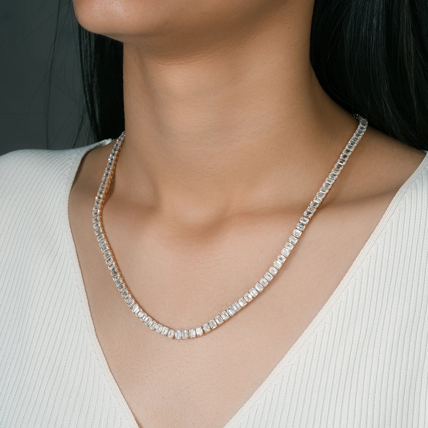 A close-up of a woman's neck and upper chest wearing a delicate tennis necklace with clear rectangular-cut stones, paired with a white ribbed V-neck top.