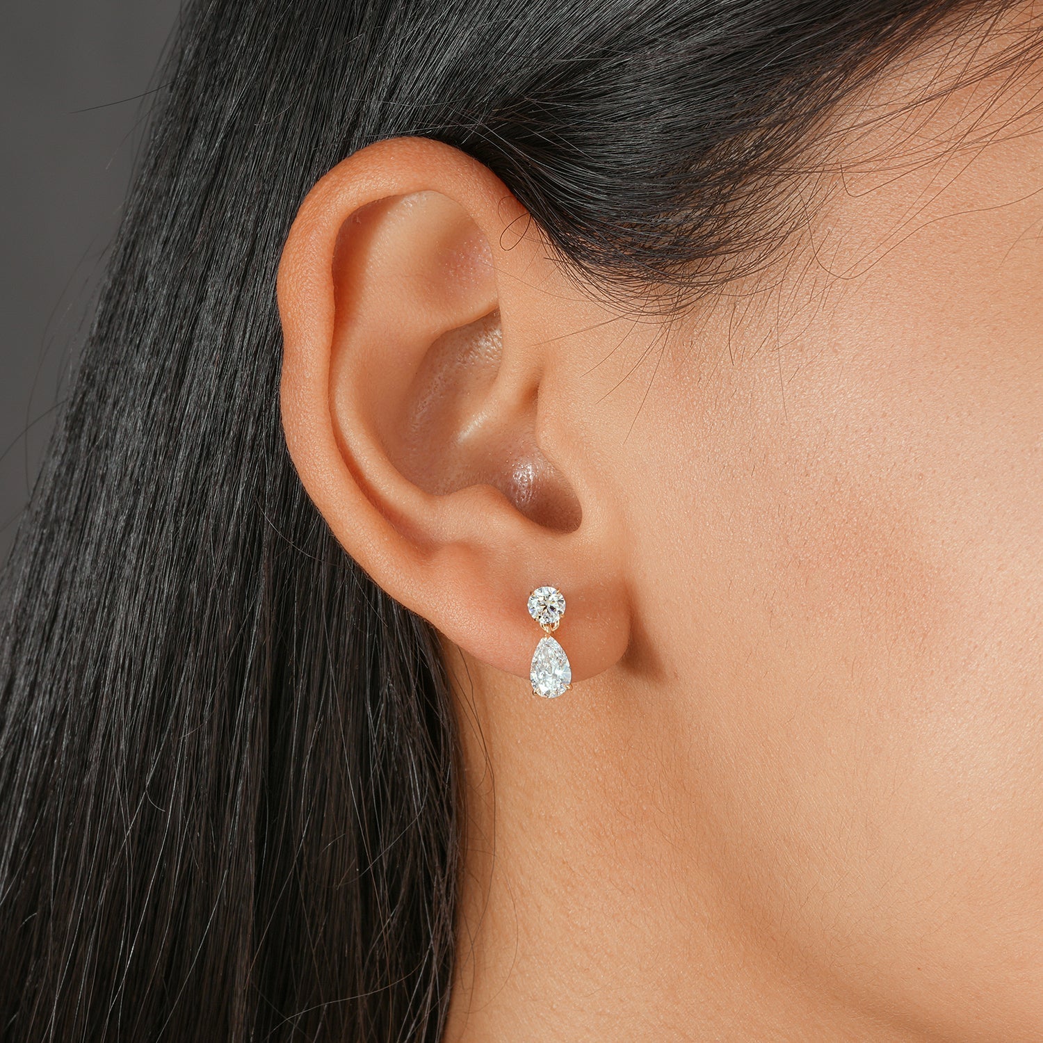 Close-up of an ear wearing a round-cut diamond with a teardrop-shaped dangling diamond earrings with a blurred background