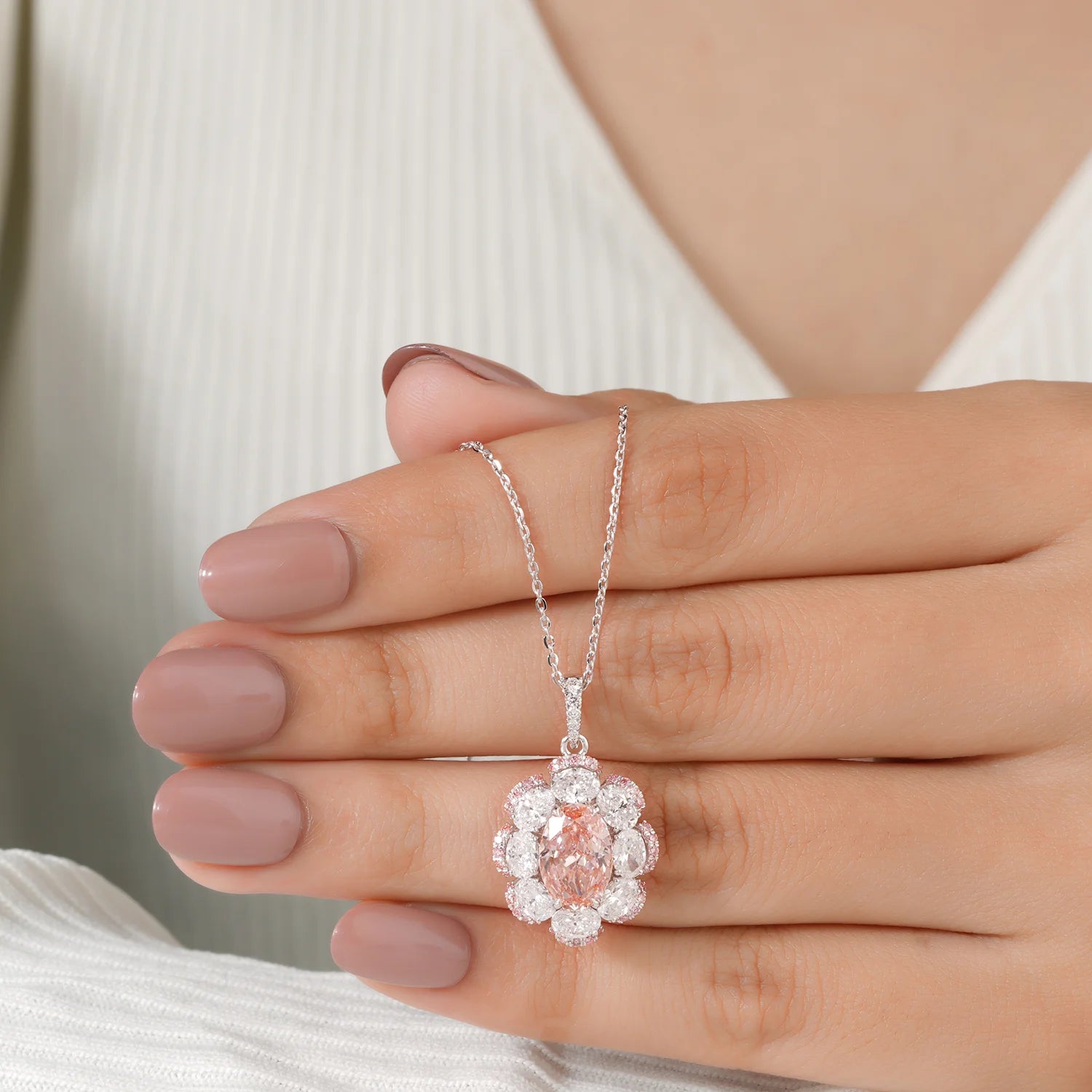 A close-up of a person's hand holding a delicate white gold pendant necklace with a central oval pink diamond surrounded by a brilliant round diamond halo, set against a soft white ribbed top.