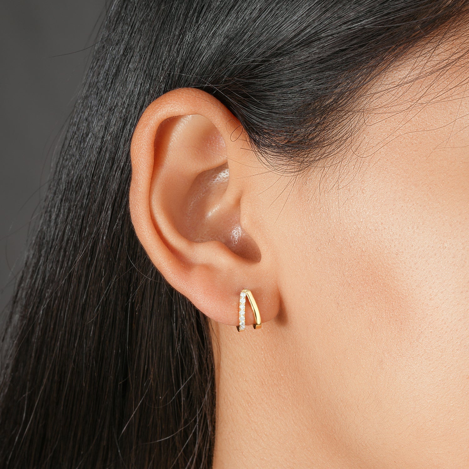 A close-up of a person's ear wearing a small curved huggie earring with a gold-toned band and a row of clear stones on one side.
