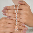 Diamond lariat necklace held between two hands with a light background. The chain is made up of different cuts of diamond, and the pendant is a blush colored pear-cut diamond.